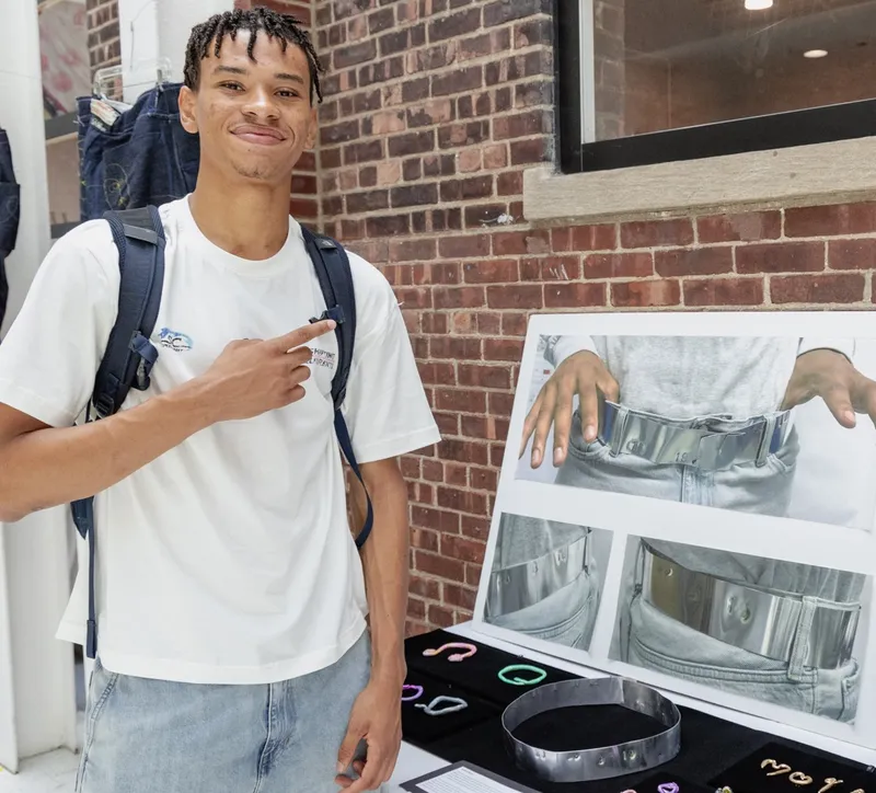 Student stands beside his art show display of a polished metal belt and small enamel jewelry.