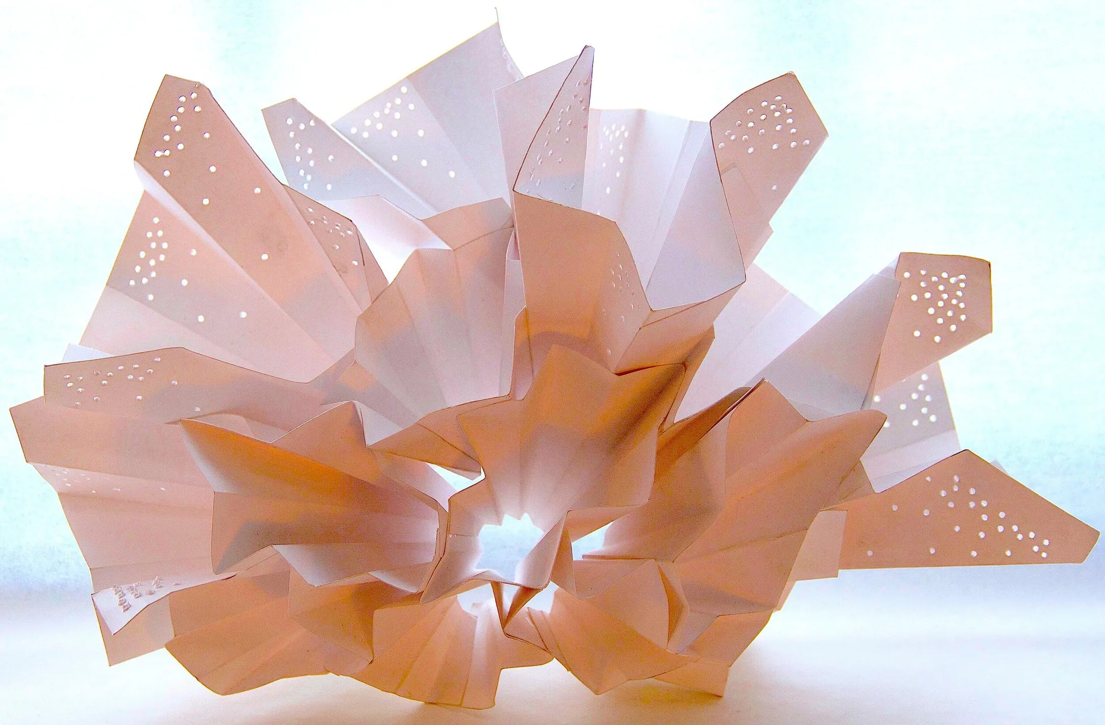 Folded white paper sculpture with pin-pricked dot patterns, lit from behind against pale blue.